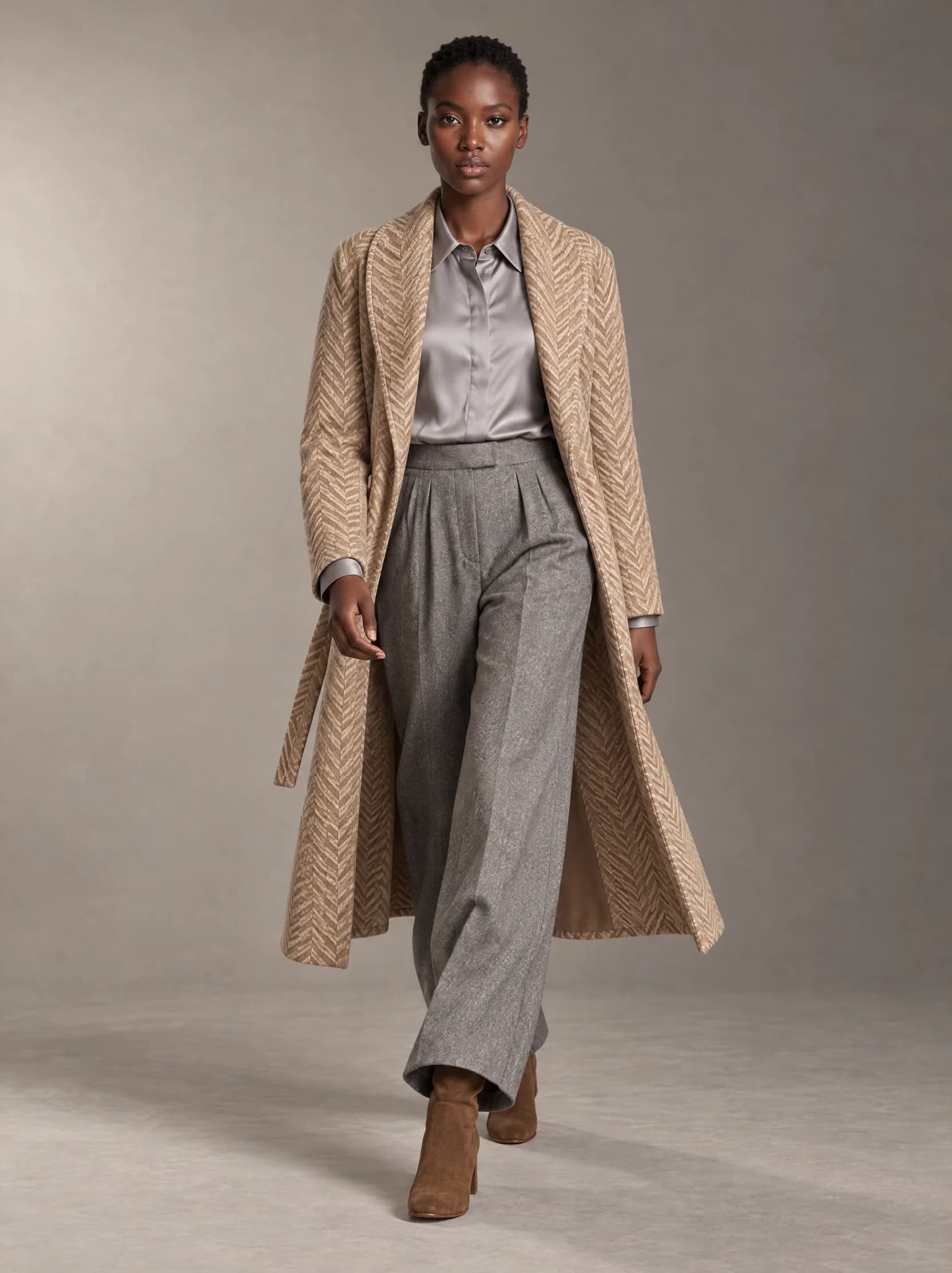 Zola walks towards the camera in the herringbone coat, worn open over a silver-grey silk shirt, with wide grey flannel trousers and brown suede boots, in a warm grey studio.