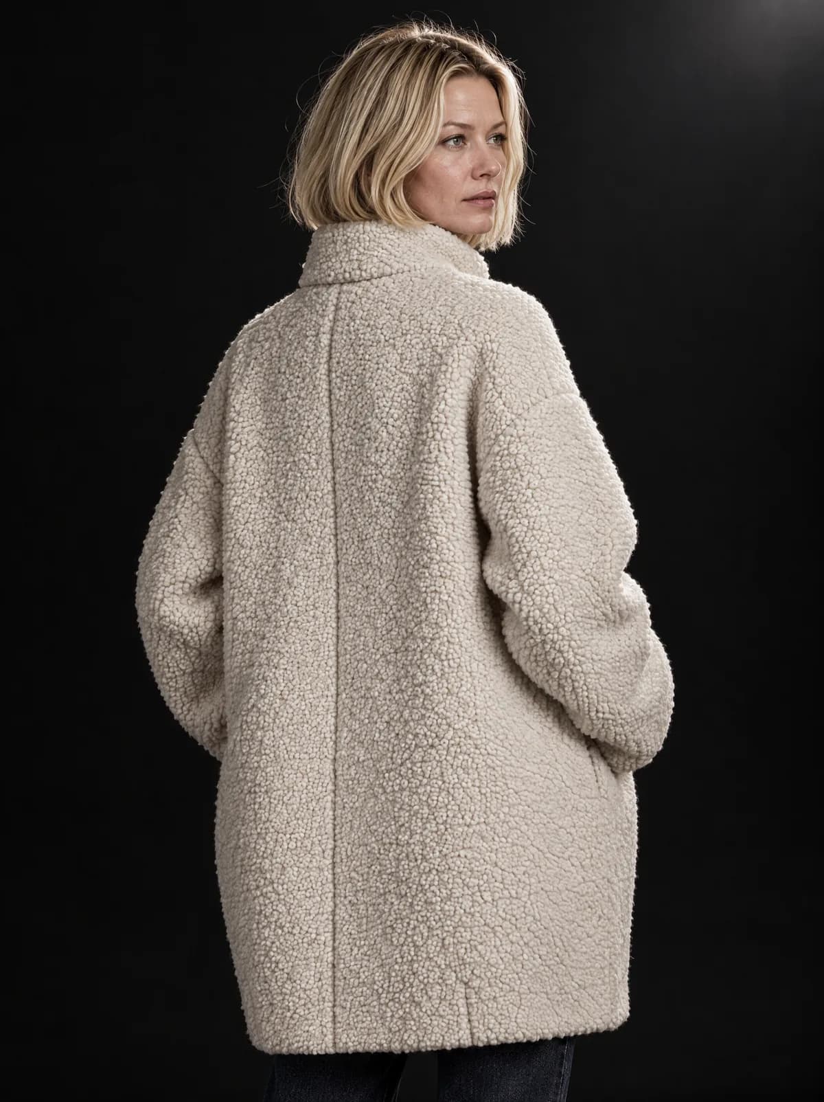 Sophie van achteren in de ivoorkleurige coconjas in bouclé; ze kijkt over haar schouder, in een zwarte studio.