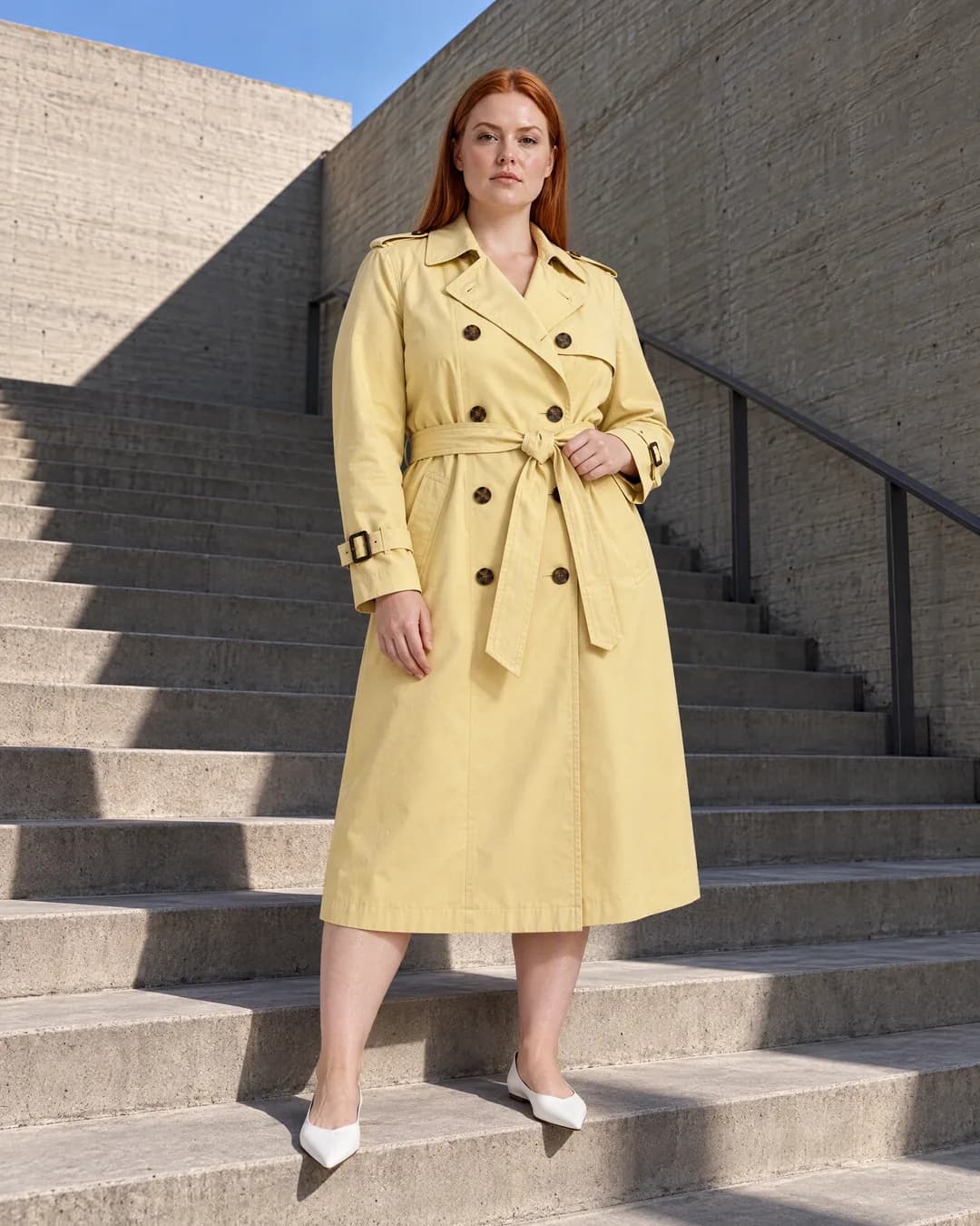 Hannah in a belted butter-yellow trench coat stands on wide concrete stairs in hard side light.